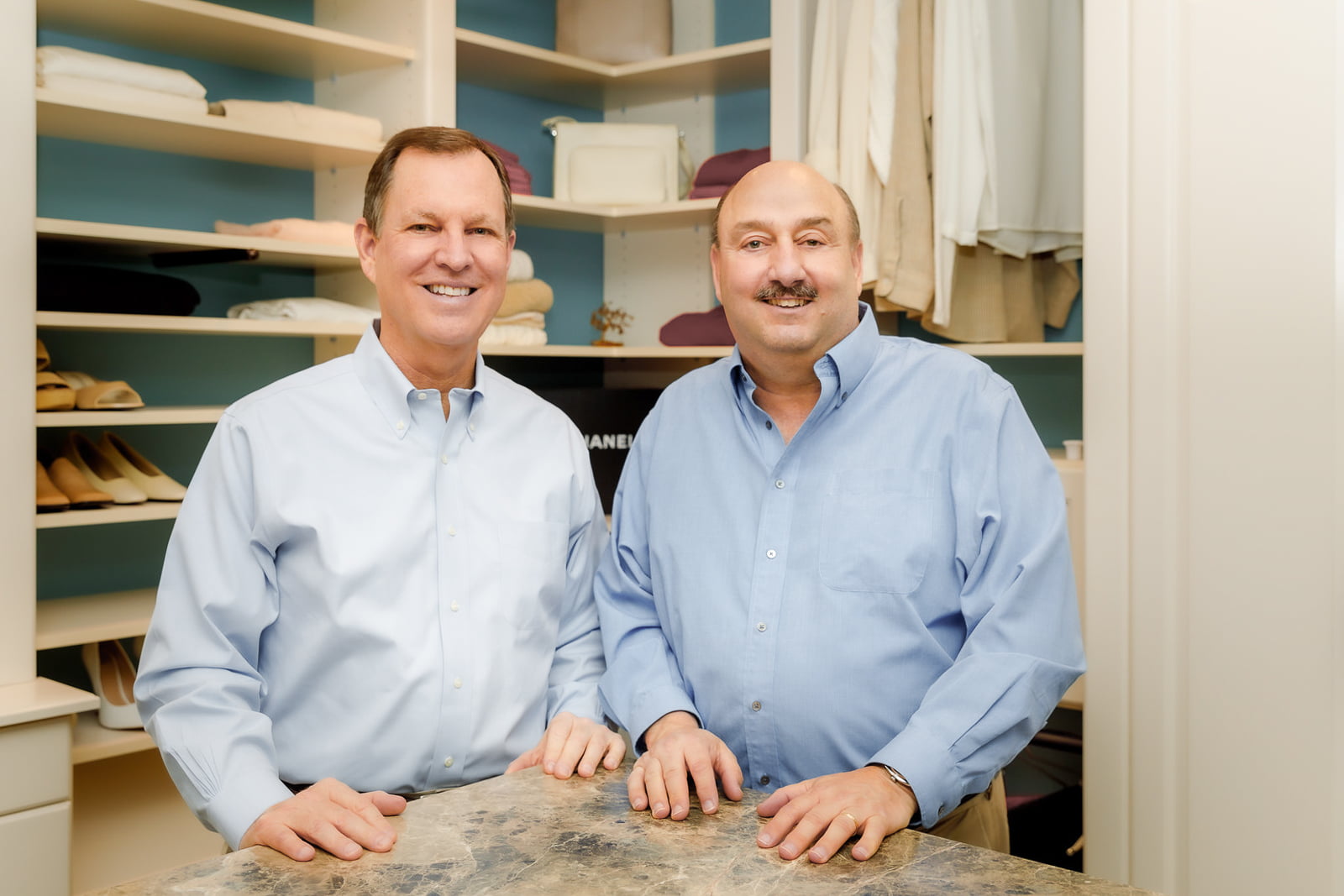 Jay and Monte in blue shirts standing in front of a closet with shelves of clothes and shoes.