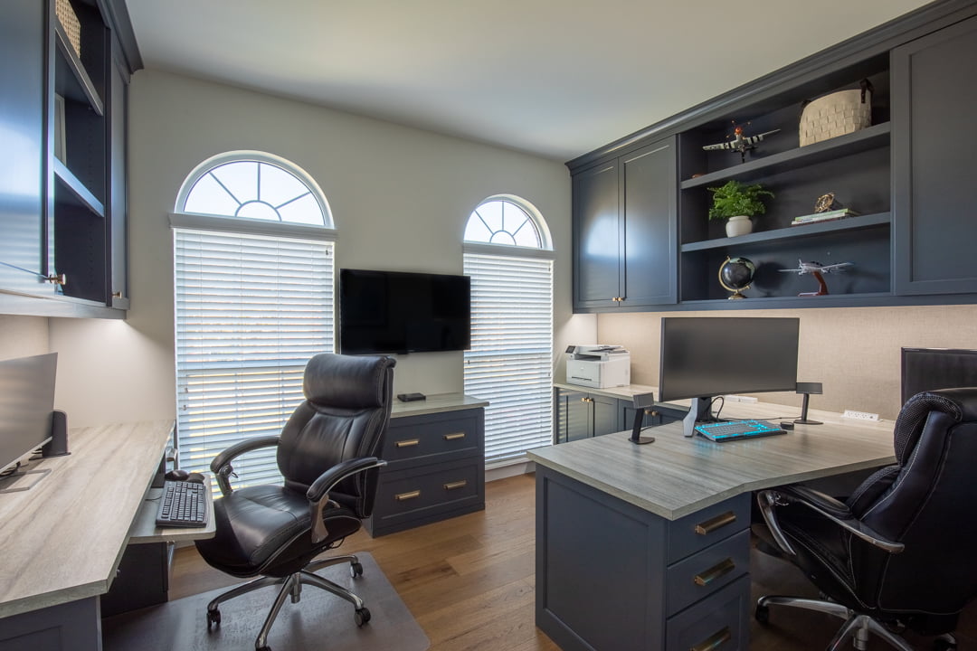 Well-lit home office with dark blue cabinets, light wood desks, dual monitors, and arched windows with blinds.