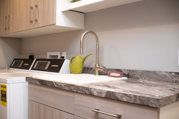 Modern laundry room with light wood cabinets, dark marble-look countertops, white sink, and stainless steel faucet.