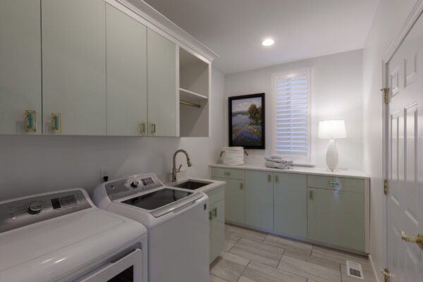Laundry room with light green cabinets, white washer and dryer, sink, window with white blinds, and a white door with brass hardware.