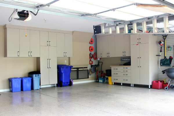 A neat, organized garage with light tan storage cabinets on beige walls, a ladder on the ceiling, and a blue trash can.