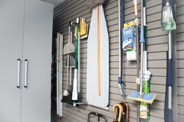 Cleaning supplies like mops, brooms, an ironing board, and dustpans hanging neatly on a gray slatwall in a utility room.