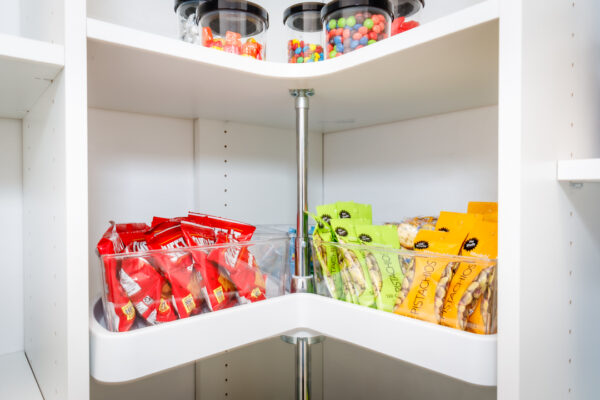 White corner pantry with a lazy Susan. Top shelf has candy jars, lower shelf has clear bins with red chip bags and green/yellow pistachio bags.