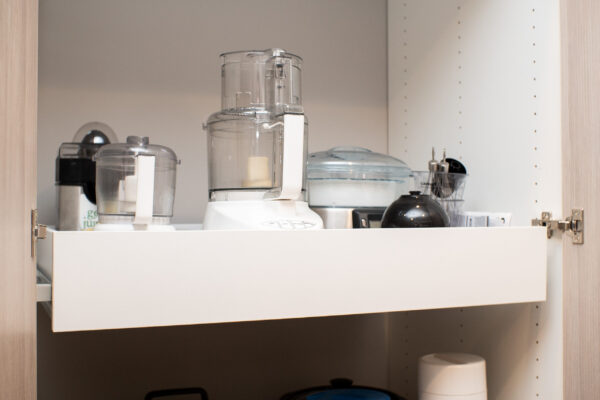 White slide-out shelf in a wooden cabinet, organized with various kitchen appliances including a food processor.
