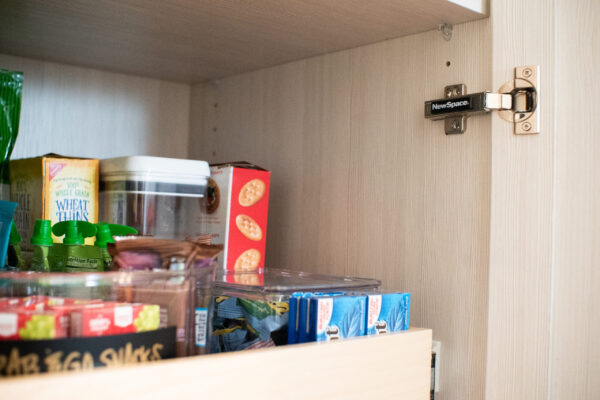 Organized pantry drawer with snacks, cracker boxes, and a "NewSpace" soft-close hinge on a light wood cabinet.