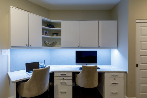 Modern L-shaped built-in home office desk with white cabinets, under-cabinet lighting, and light blue backsplashes.