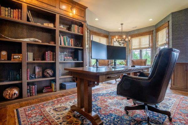 Luxurious traditional home office with large wooden desk, dual monitors, leather chair, and built-in bookshelves.