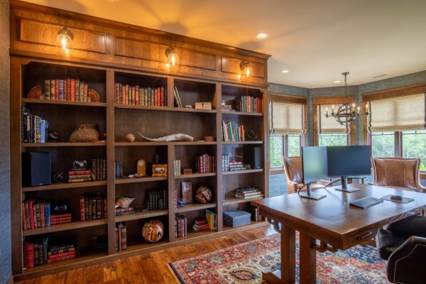 Rustic home office with large wooden bookcase, ornate rug, and two monitors on a desk.