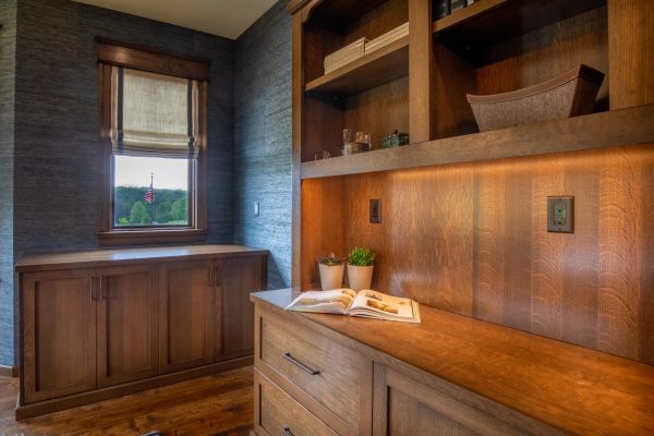 Home office with dark gray textured wallpaper, custom wood cabinetry, a built-in desk, and a window with a Roman shade and an American flag outside.