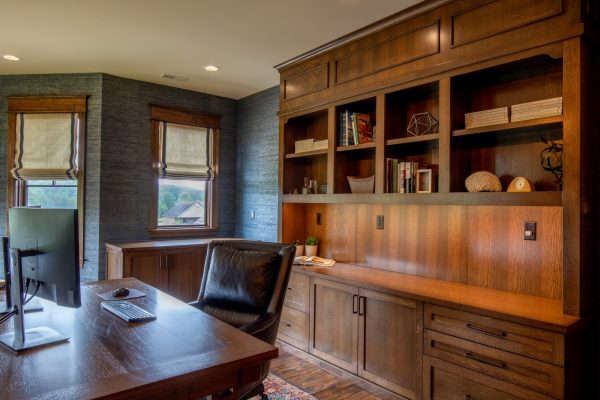 A luxurious home office with dark wood desk, black leather chair, built-in wooden shelves, and gray textured walls.