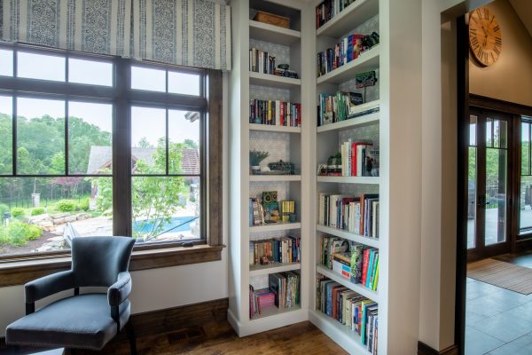 Built-in white bookshelves filled with books next to a large window overlooking a lush green yard and pool.