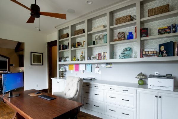 A home office with white built-in shelves featuring geometric patterns, books, and decor, a dark wooden desk with a computer, and a light-colored armchair.