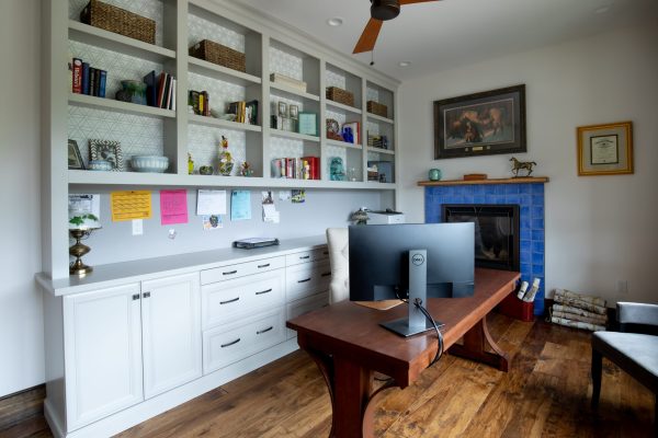 A cozy home office with gray built-in shelves and white cupboards. A dark wood desk with a monitor sits before a blue-tiled fireplace.