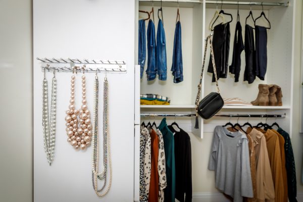A white closet with necklaces hanging on the left and various clothes, boots, and a handbag on shelves and hangers.