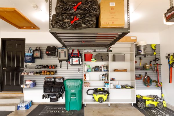 An organized garage with a black door, hanging shoes, bags, tools, shelves, and a yellow lawnmower.