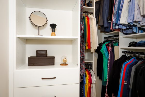 Walk-in closet with white shelves storing mirror, grooming brush, brown boxes, and gold dome clock, beside hanging clothes.