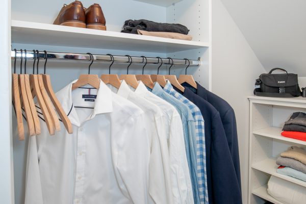 White walk-in closet with shelves, hanging shirts, brown shoes, and a wicker basket on a light wood floor.