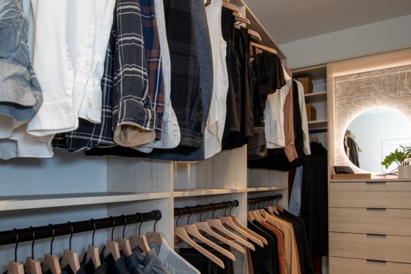 Modern walk-in closet with hanging clothes, light wood shelving, and a dresser with an illuminated round mirror.