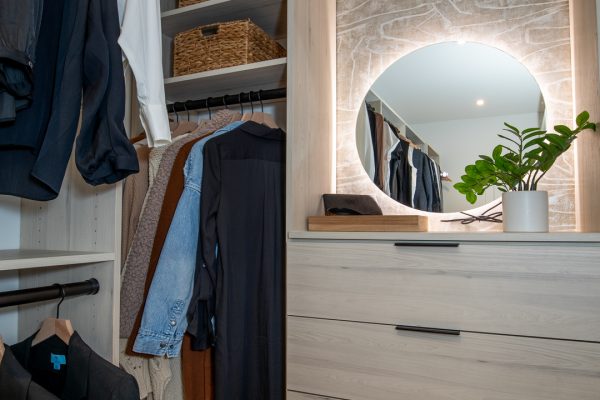 Modern light wood closet system with hanging clothes, wicker baskets, and a dresser with a round lighted mirror.