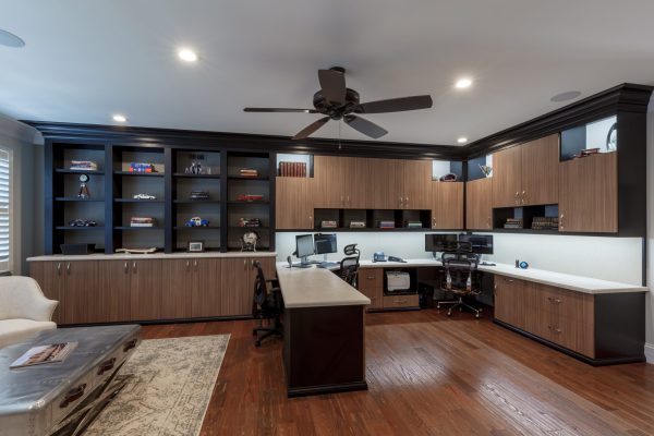 Spacious home office with dark wood floor, modern wood cabinetry, black shelves, multiple computer workstations, and a ceiling fan.