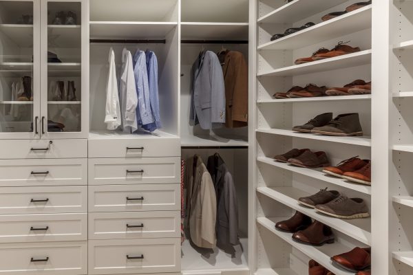 Custom walk-in closet: white drawers, glass cabinets with boots, hanging shirts, jackets, & multiple shoe racks.