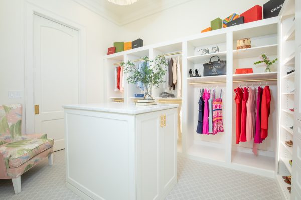 Luxurious white walk-in closet with central island, organized clothing, designer bags, and patterned chair.