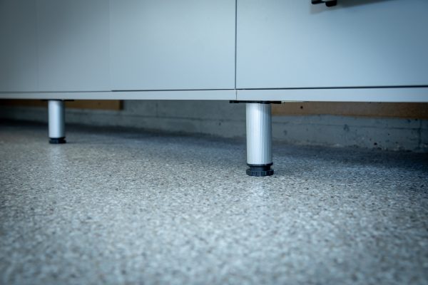Metallic ribbed leveling leg supports a modern gray cabinet on a speckled floor.