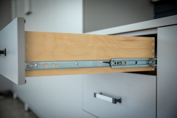 Open wooden drawer with a light gray face and silver metal slides, extending from a white cabinet.