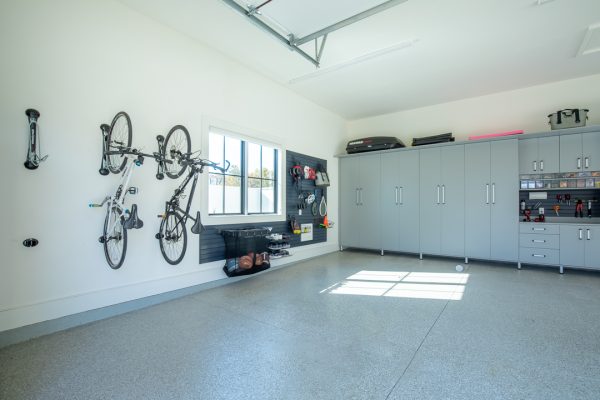 Bright white garage with two bikes hanging, a window, black slatwall organizing tools, and grey storage cabinets.