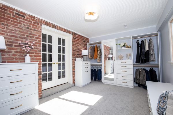Luxurious walk-in closet with exposed brick, white dressers, French doors, & built-in shelving with hanging clothes.