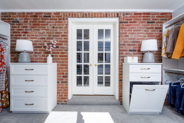 Custom closet organization system in a room with exposed brick walls, white French doors, and two white dressers with gold handles.