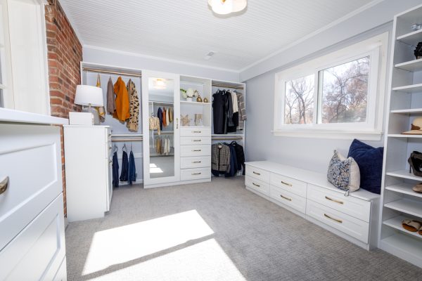 Spacious custom closet with white built-ins, clothes on racks, drawers, window, and a brick wall.