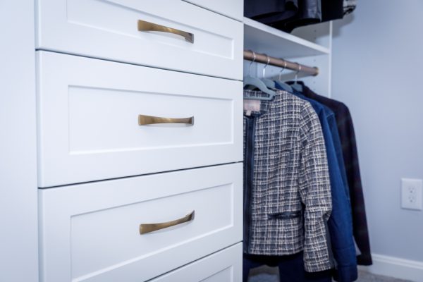 Custom white closet drawers with modern brass handles next to hanging clothes.