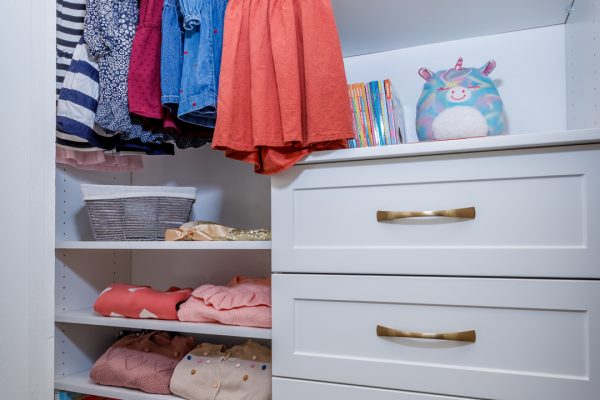 Children's white closet with dresses, folded clothes, books, and drawers with gold handles.