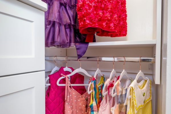 Custom white closet with colorful children's party dresses, casual clothes, and vests hanging on metal rods.