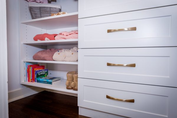 White custom closet with shelves holding folded clothes, children's books, a basket, and drawers with gold handles.