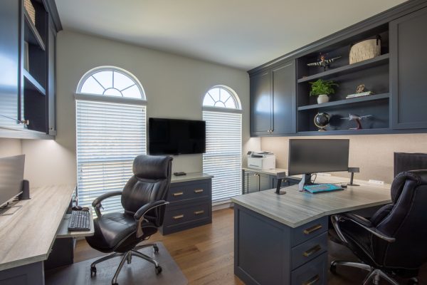 A spacious home office with custom navy cabinets, light wood desks, two black desk chairs, dual arched windows, and decorative shelves.