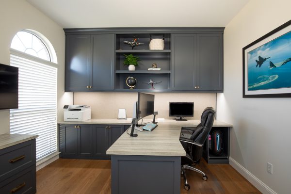 Modern home office with dark blue built-in cabinets and light wood desk, window, and fighter jet art.