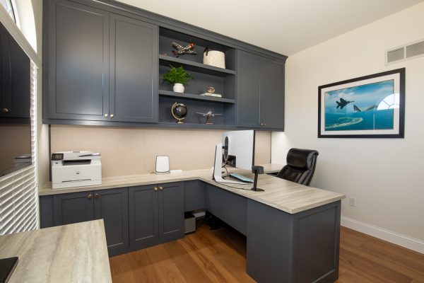 Modern home office with dark gray built-in cabinets, a light wood U-shaped desk, a printer, and a monitor.