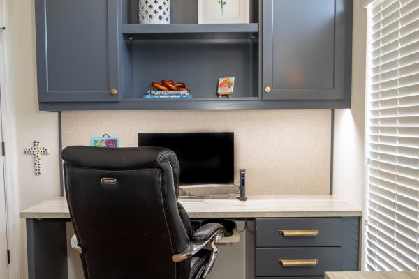 Built-in gray home office desk with black ergonomic chair, open shelving, and light wood flooring.
