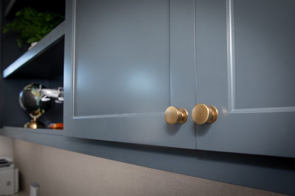 Close-up of matte gray cabinets with round brass knobs, an open shelf with a plant and globe in the background.