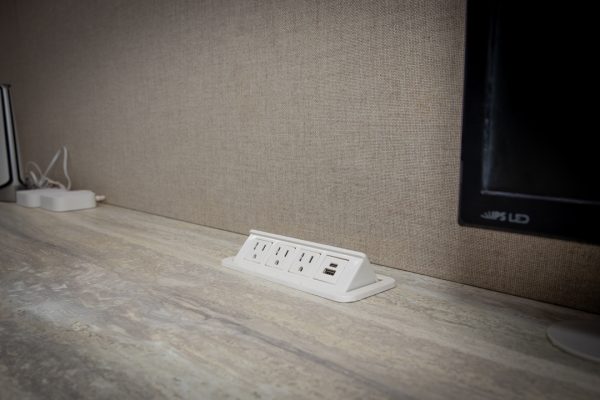 White power strip with three outlets and two USB ports, recessed into a light gray wood-grain desk.