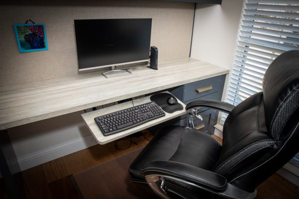 Modern home office desk with light wood-grain top, black monitor, keyboard tray, and black executive chair.